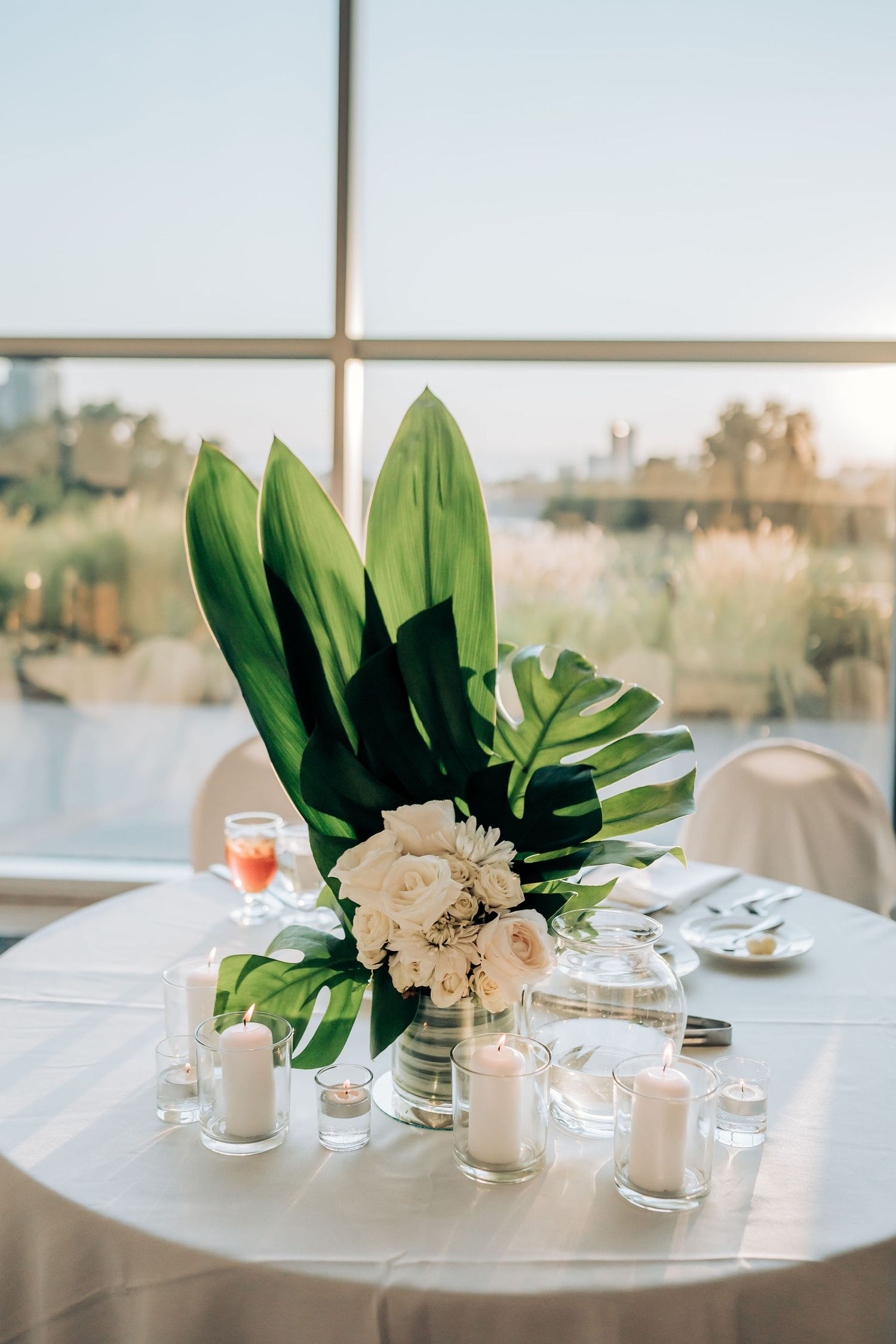 Modern wedding centerpiece with tropical leaves and white roses on round table by window.