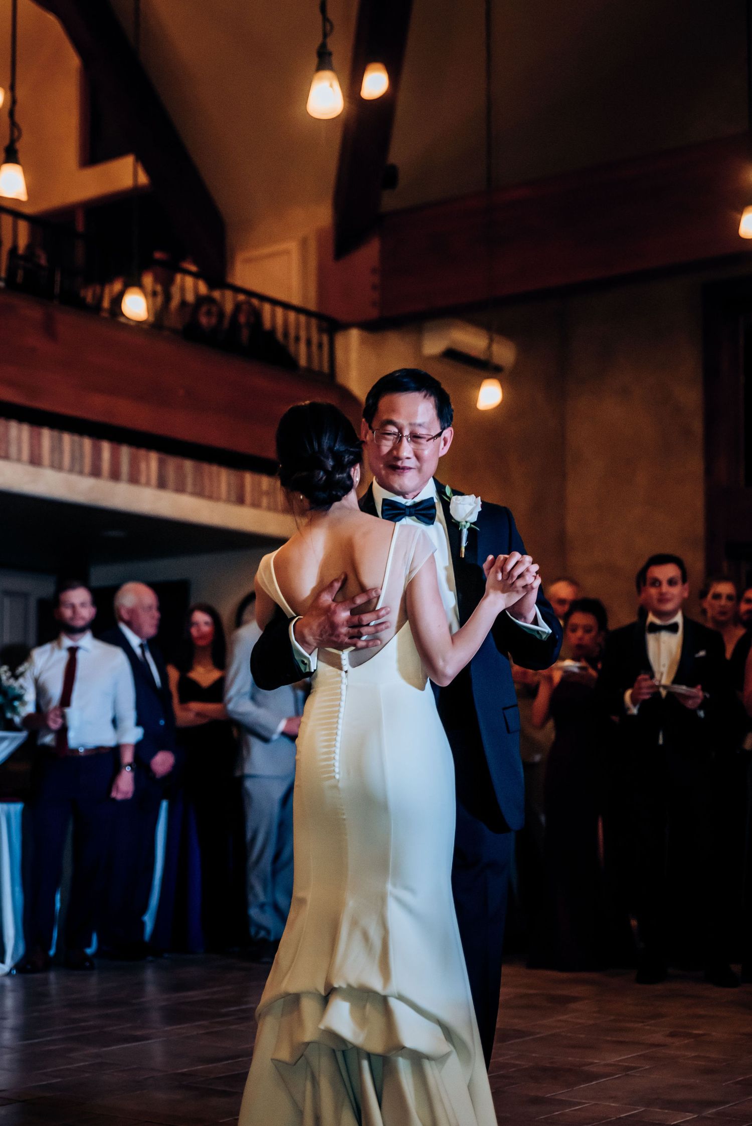 Father daughter dance at a wedding reception in a rustic venue with warm lighting and watching guests.