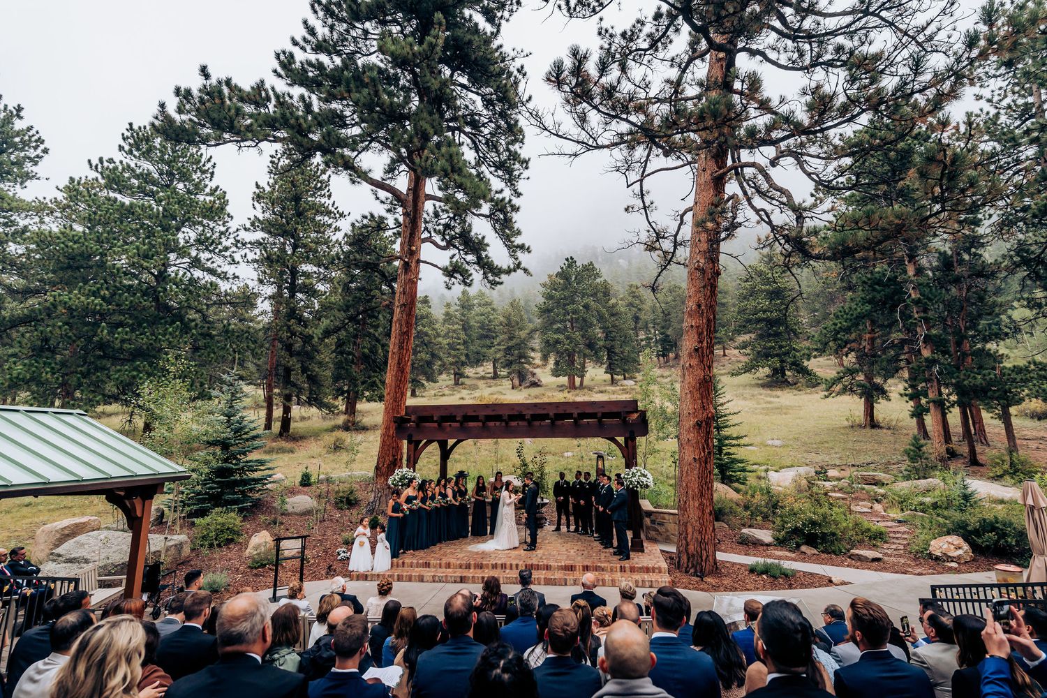 A mountain wedding ceremony viewed from above showing guests seated before a wooden pavilion among pine trees.