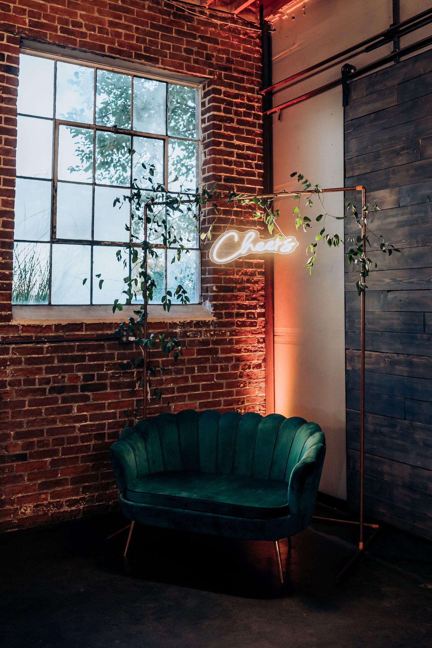 Modern industrial venue space with emerald velvet couch and neon sign against exposed brick wall.