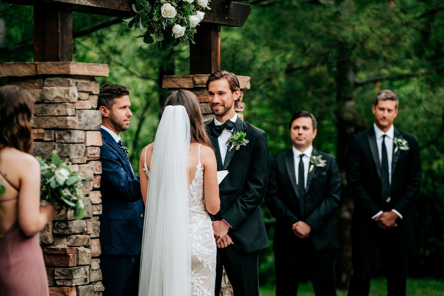 A wedding ceremony takes place outdoors under a stone archway with greenery in the background.