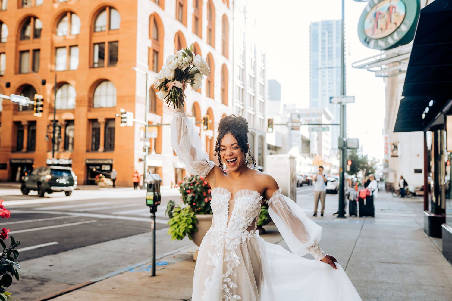 Elegant bride in white dress celebrates on urban street with historic brick building in background.