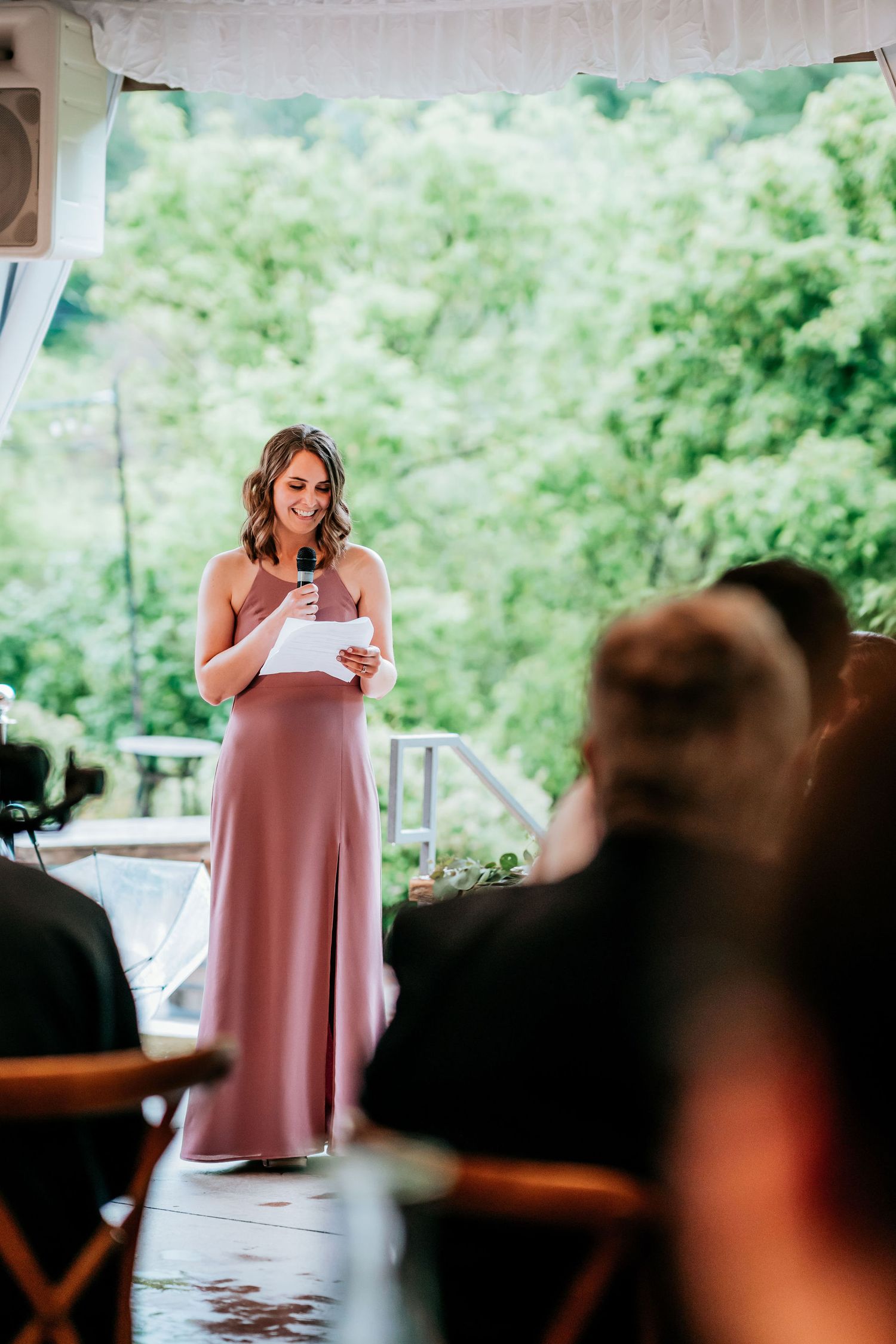 A bridesmaid in a mauve dress gives an emotional speech during a wedding reception on an outdoor porch.