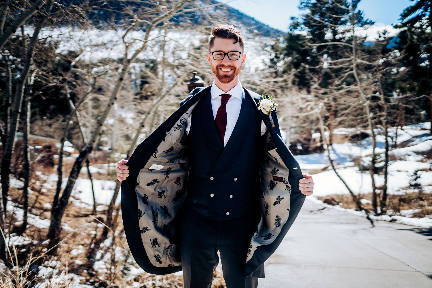 Groom in navy suit poses playfully with jacket in snowy mountain setting.