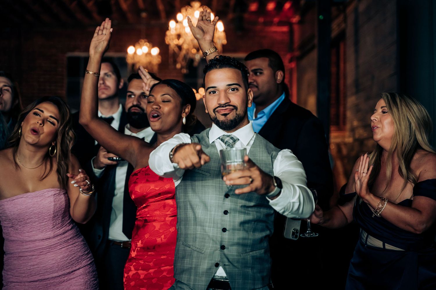 Party guests dance and celebrate beneath crystal chandeliers at an indoor venue.