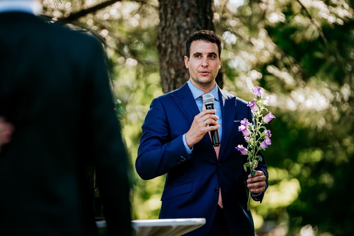 A person in a navy suit speaks into a microphone while holding flowers at an outdoor event.