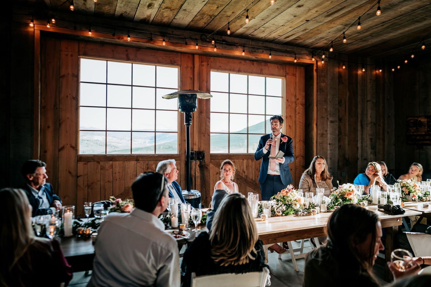 Wedding speaker addresses guests in a rustic wooden venue with string lights and large windows.