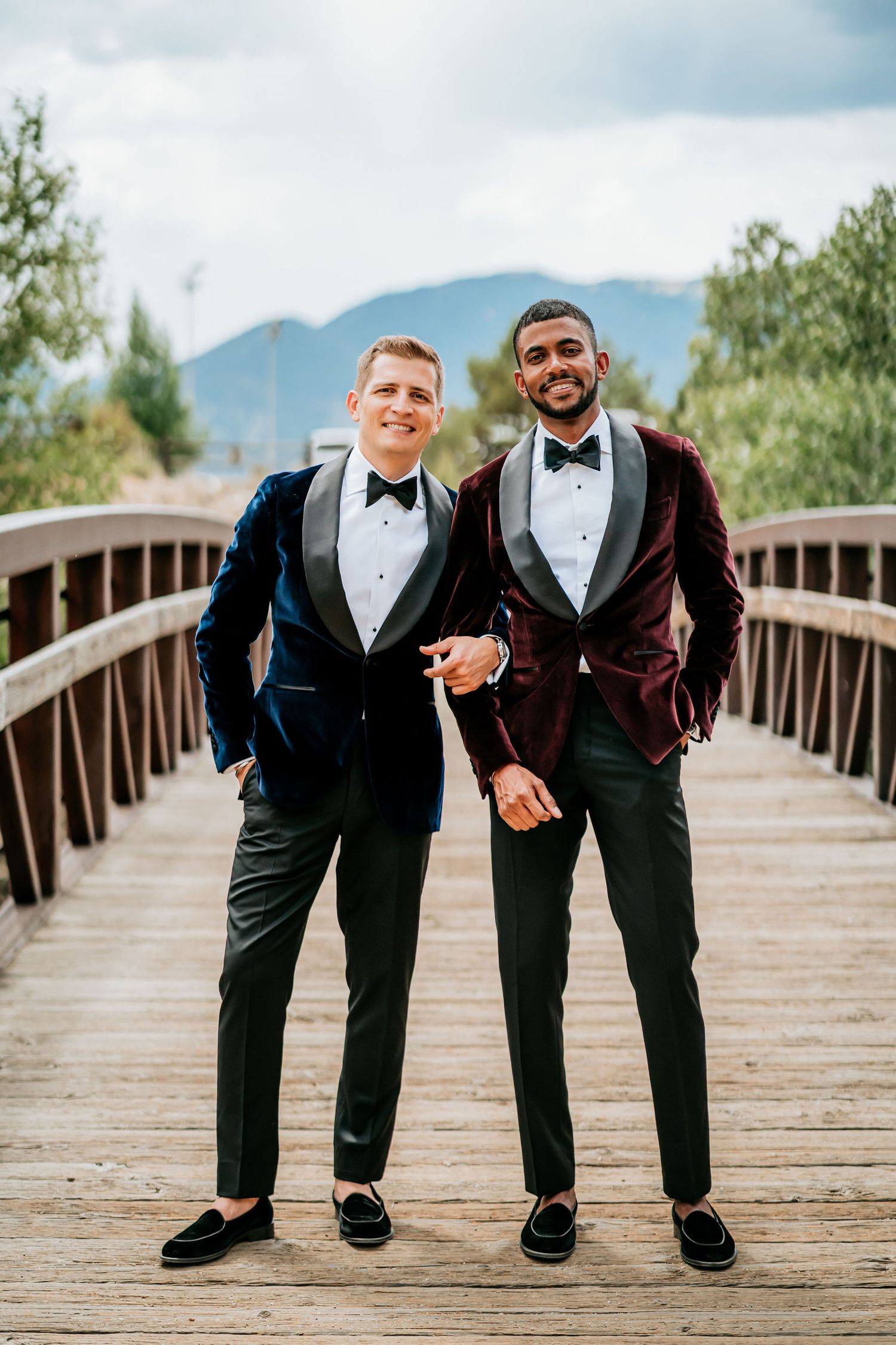 Couple in formal tuxedos standing together on a wooden bridge with mountains in background.