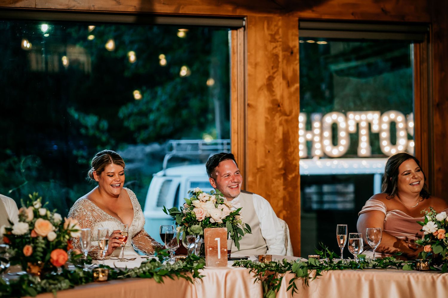 Wedding reception guests seated at a decorated head table with string lights and wood accents.