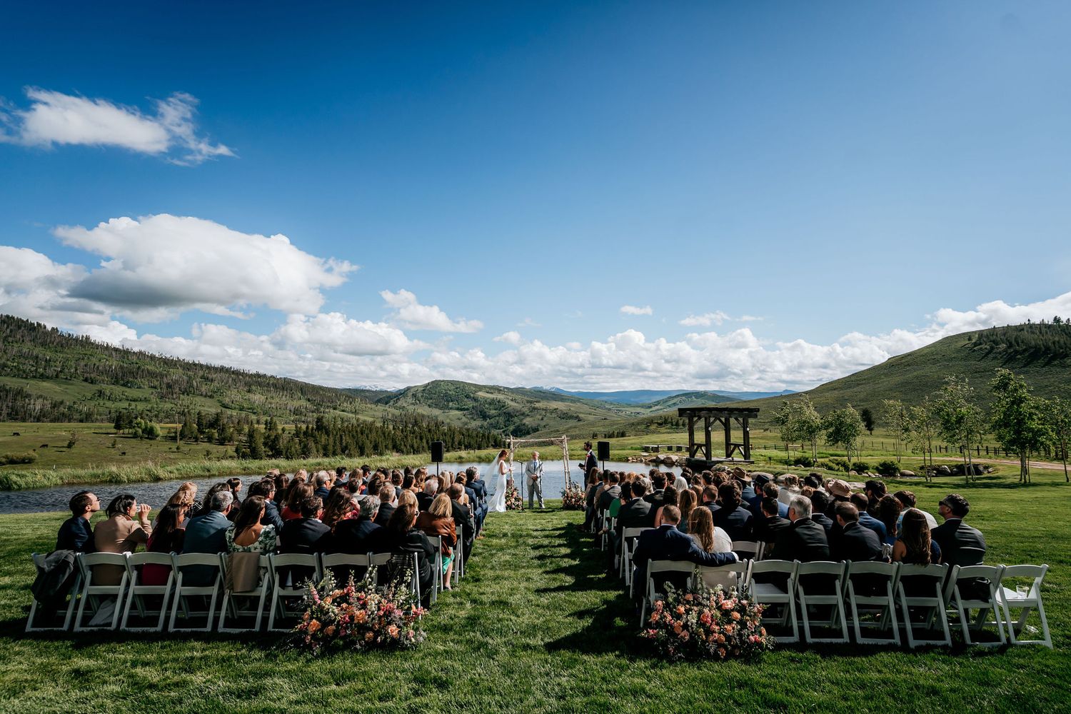 Outdoor wedding ceremony setup with white chairs arranged in rows facing mountains and lake under blue skies.