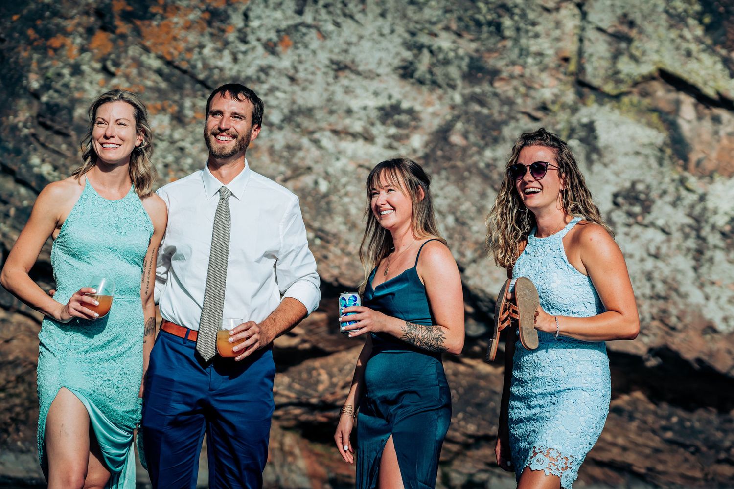 Wedding party members in blue attire enjoy drinks by a rocky cliff backdrop.