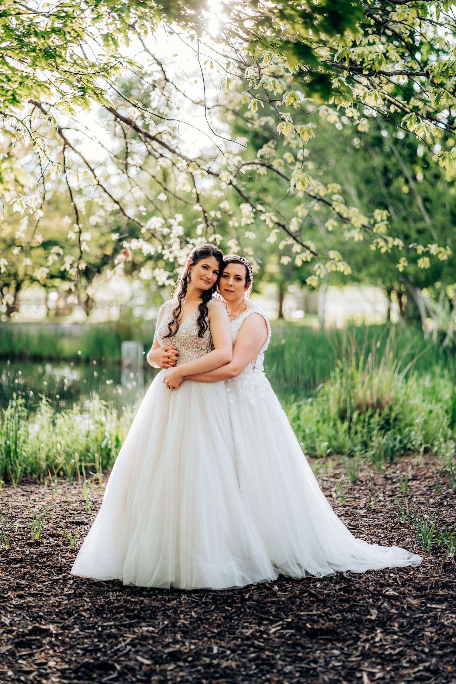 Couple in matching white wedding dresses poses together under sunlit tree branches.