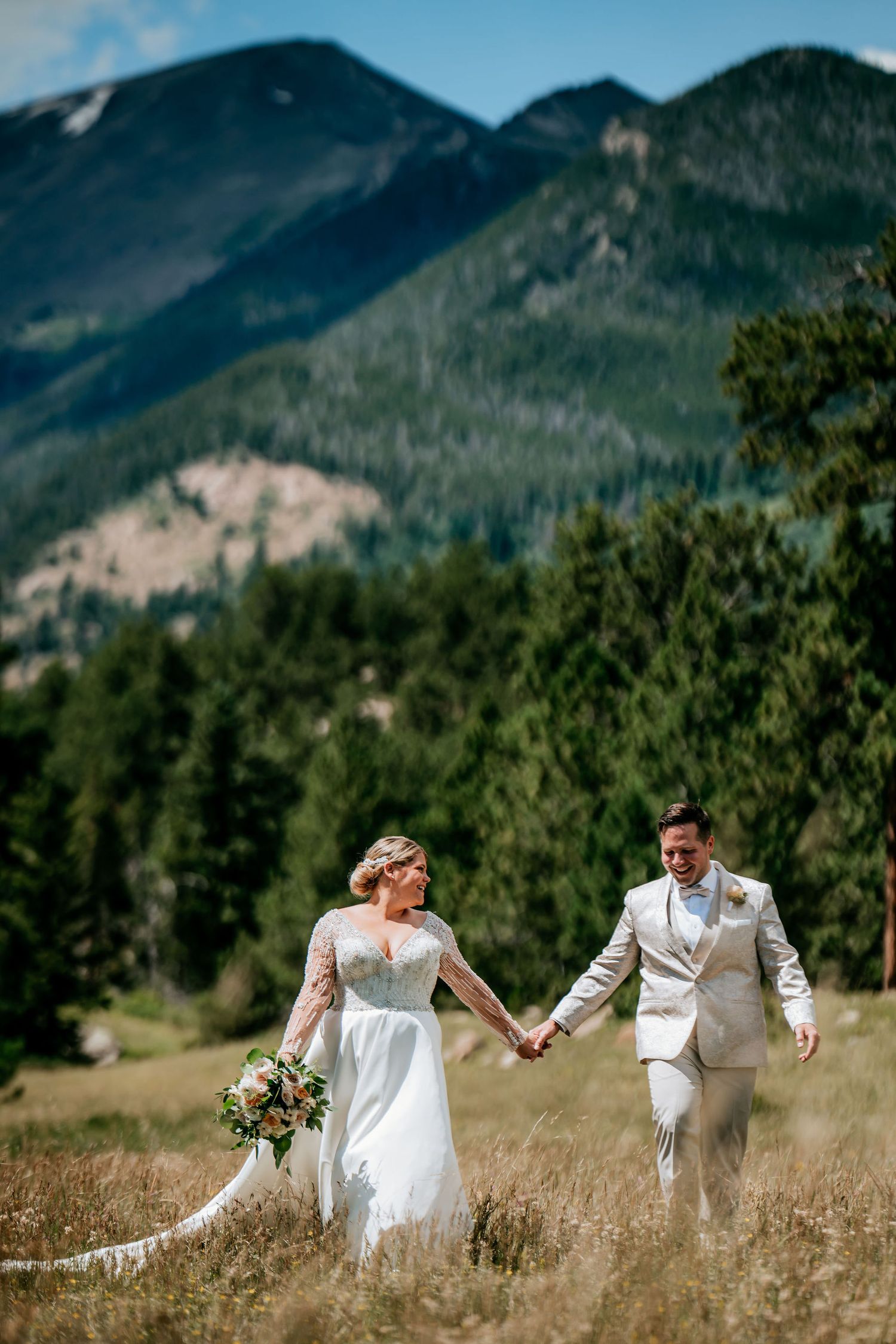 Couple running hand in hand through mountain meadow with dramatic peaks in background.
