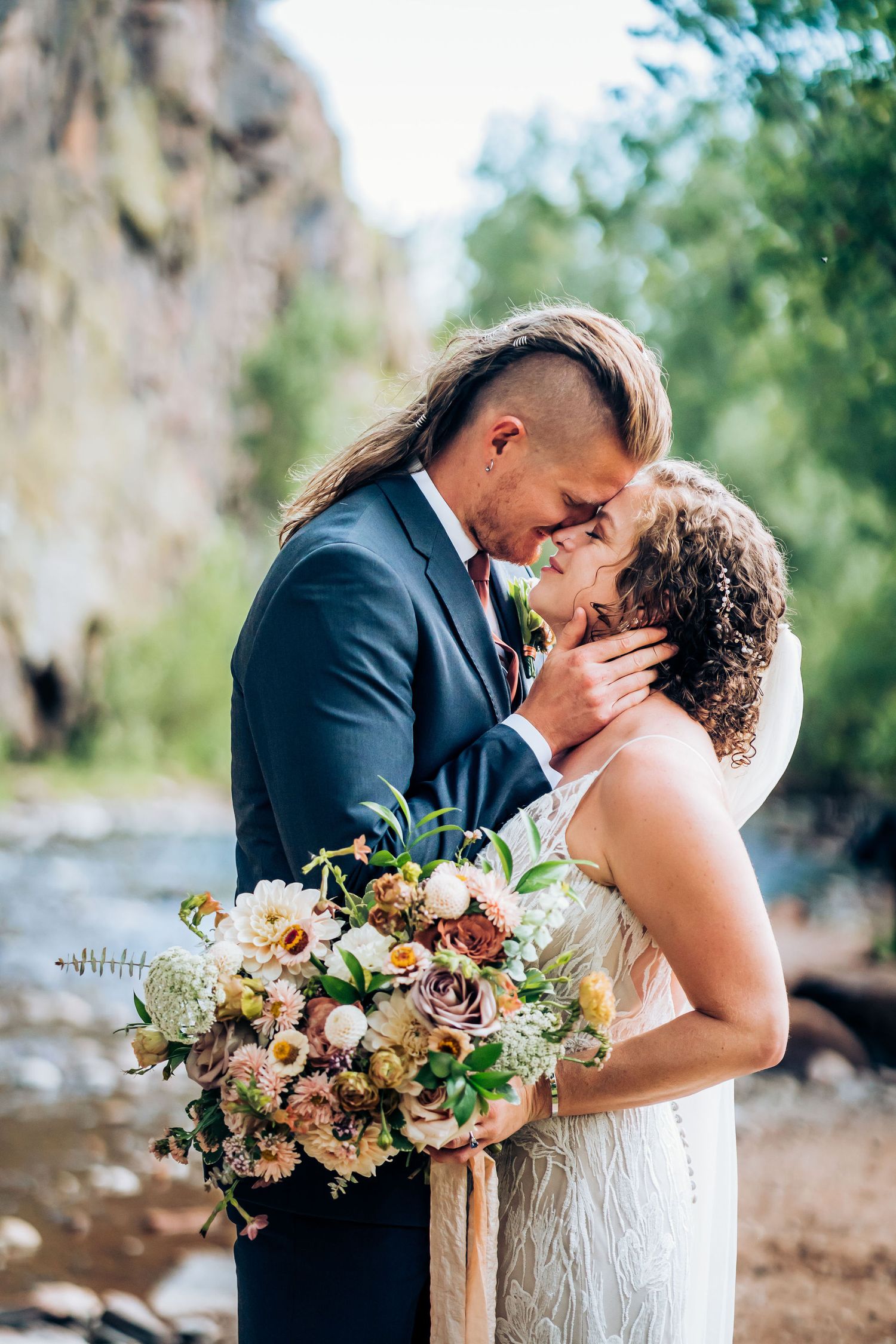 Tender moment between newlyweds with lush floral bouquet against rocky natural setting.