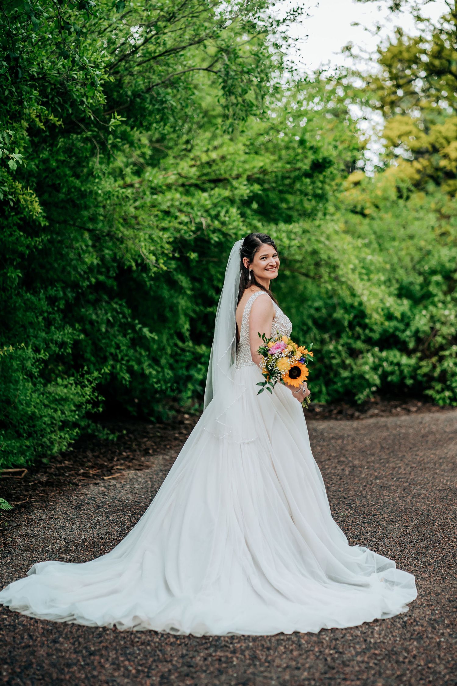 Bride in flowing white gown holding sunflowers poses on garden path with trailing veil.