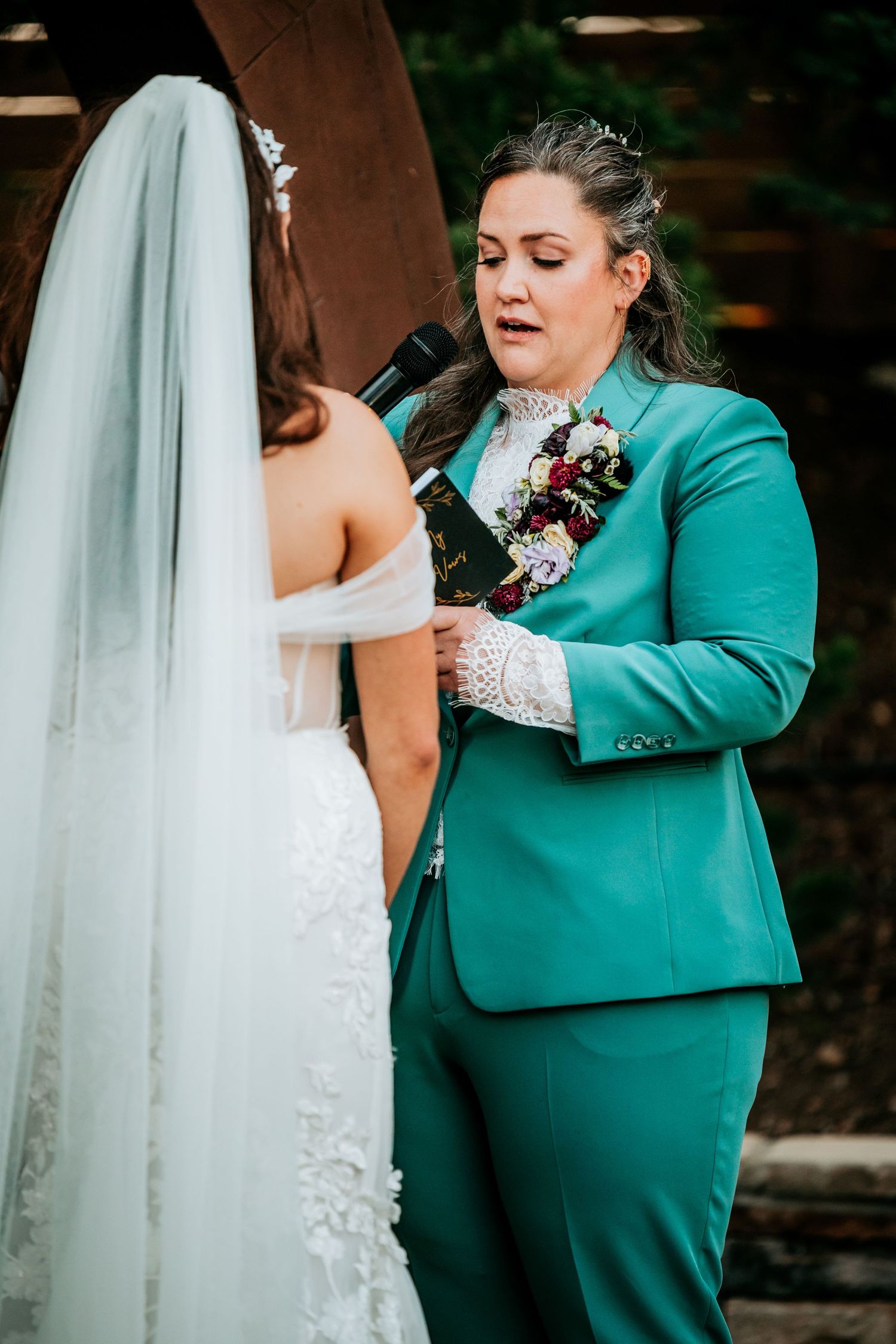 Partner in elegant teal suit delivering wedding vows at outdoor ceremony beneath floral arch.