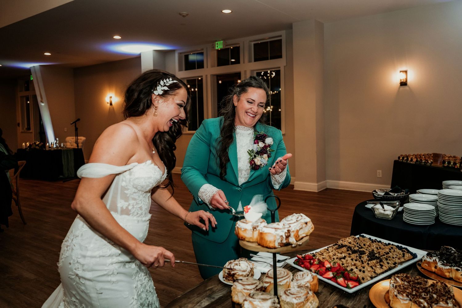 Two brides smile and cut their wedding cake together at a reception dessert table with assorted treats.