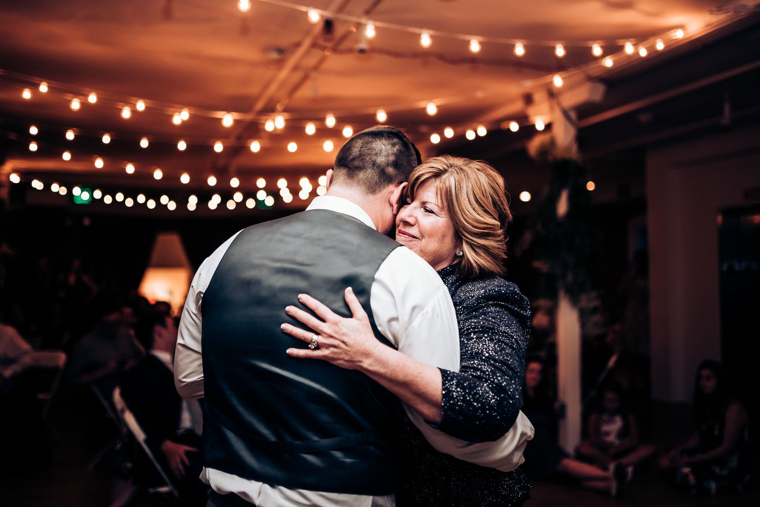 Two people share an intimate dance moment under glowing string lights at an indoor celebration.
