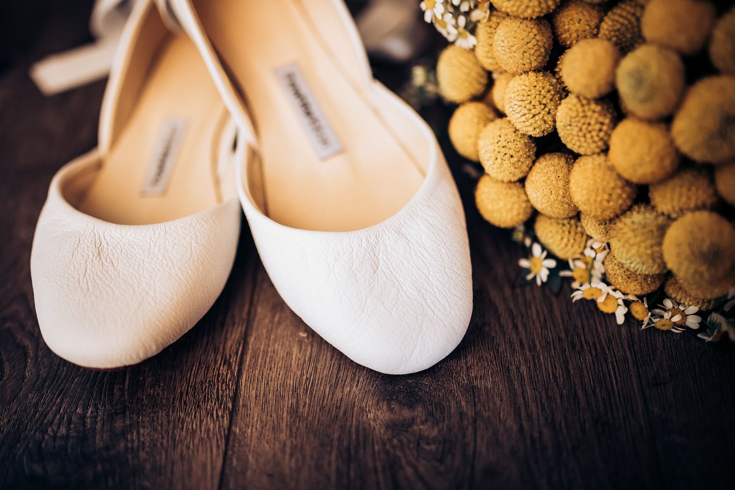 Delicate white ballet flats and yellow billy ball flowers arranged on dark wooden surface.