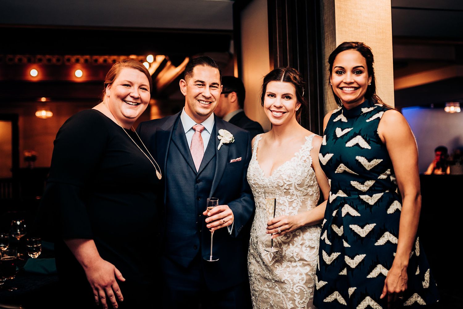 Four guests smile and pose together at an indoor wedding reception with warm lighting.