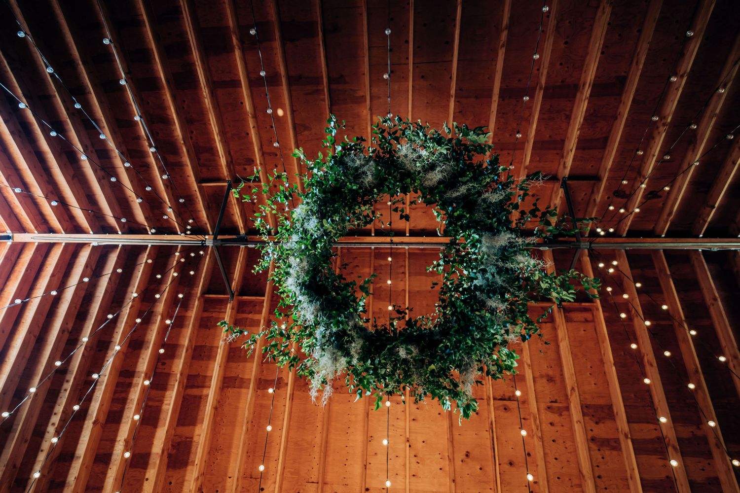 Green holiday wreath hanging from wooden beams with twinkling string lights.