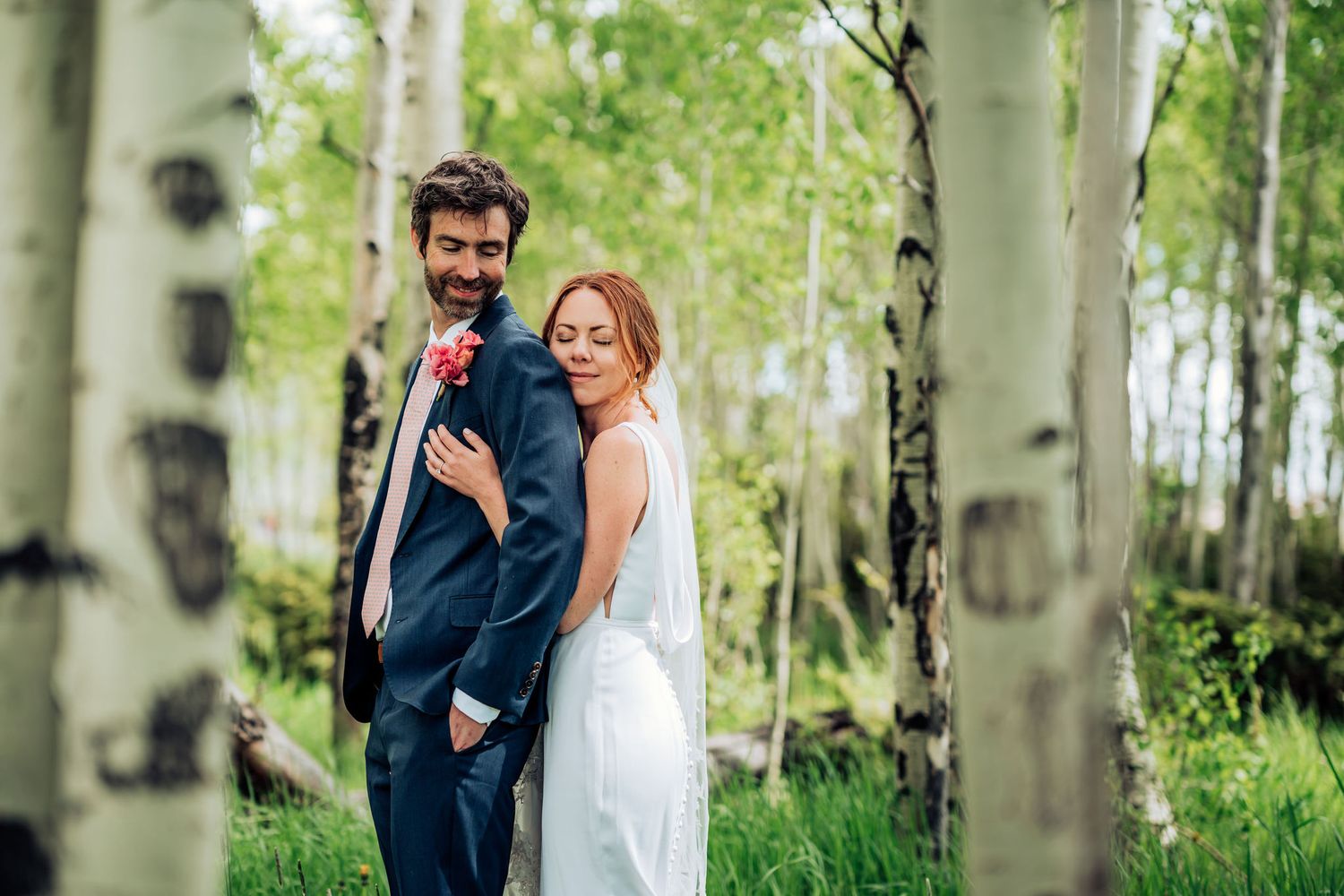 Joyful wedding couple poses playfully between white birch trees in a sunlit forest.