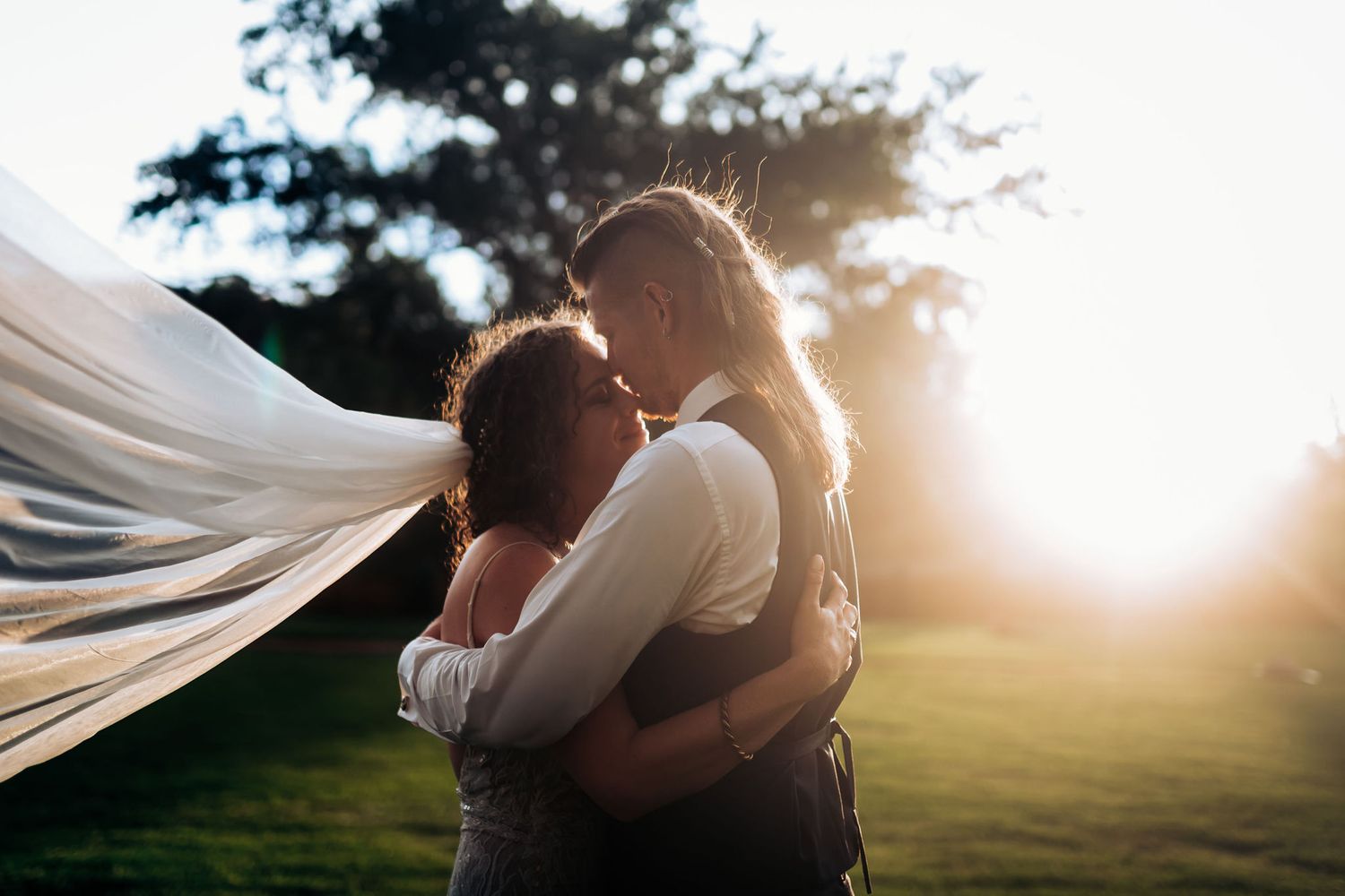Silhouetted couple embraces during sunset with flowing veil in breeze.