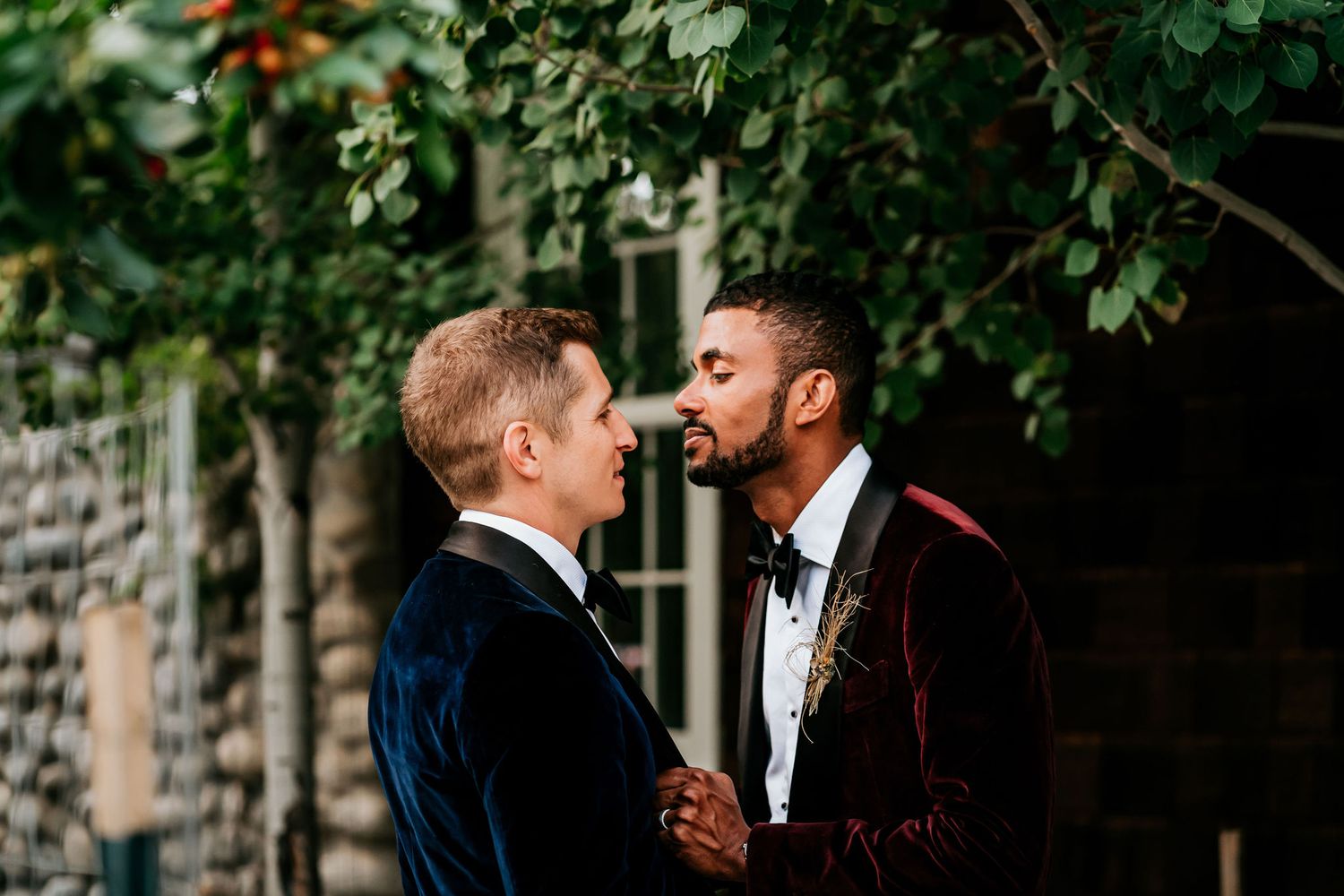 Two people in tuxedos share a tender moment against a stone wall backdrop.