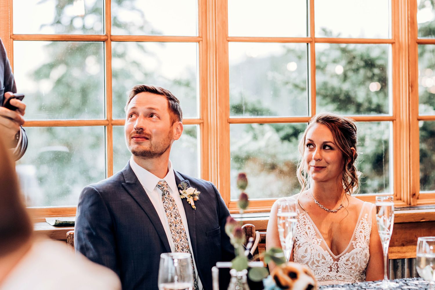 Couple seated at head table during wedding reception near bright windows.