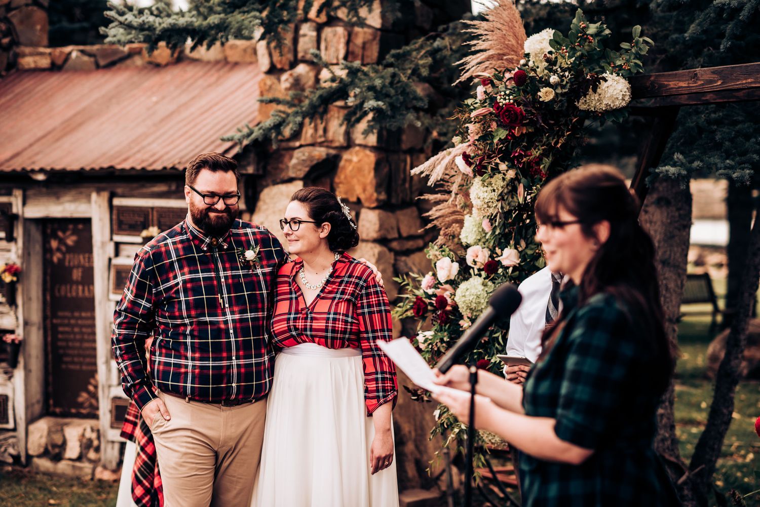 Wedding participants in coordinating plaid shirts gather near a floral arch for an outdoor ceremony.