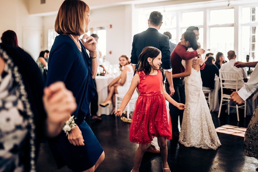 Young girl in a red dress dances at a wedding reception while guests celebrate nearby.