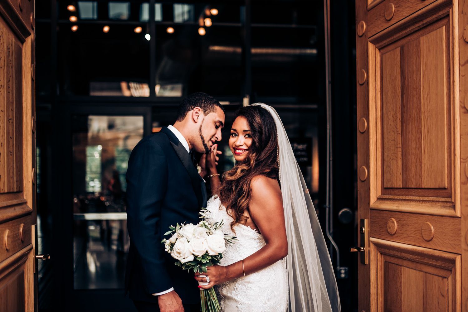 A romantic wedding moment captured in front of grand wooden doors as couple holds white flowers.