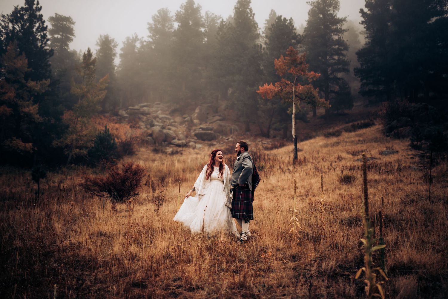 Wedding photo of couple in traditional dress on misty hillside with evergreen forest backdrop.