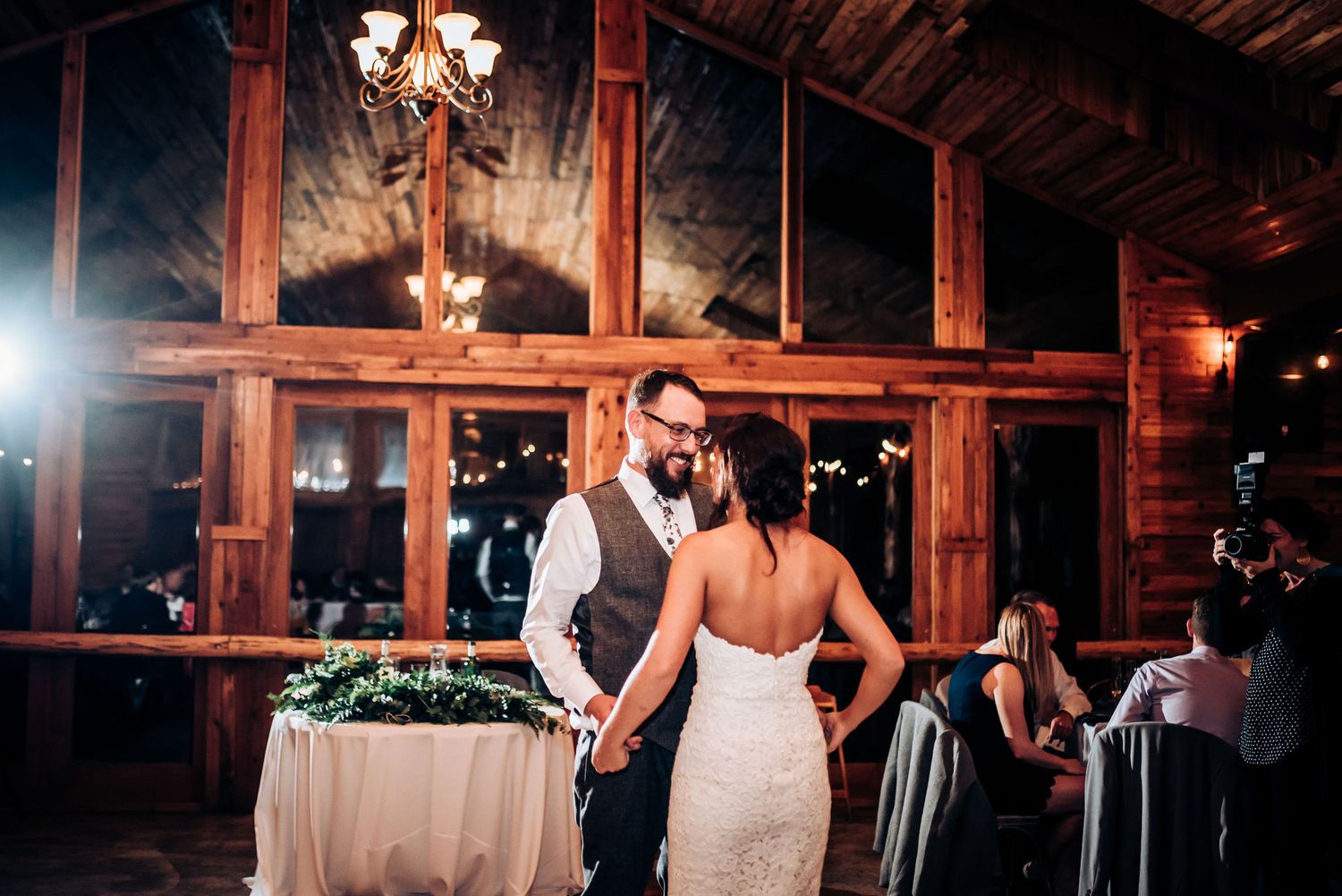 Couple dances near decorated tables in rustic barn venue with large windows.