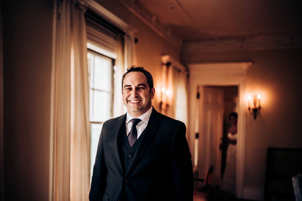A groom in a dark suit stands in an elegant hallway with wall sconces.