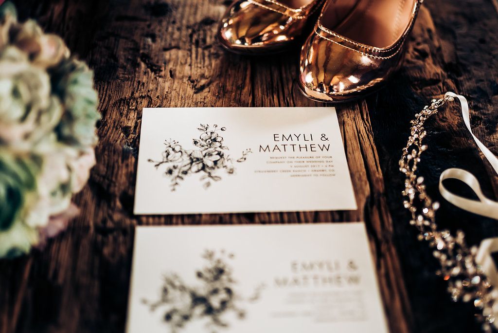 Floral wedding invitations with metallic shoes and delicate accessories on wooden surface.