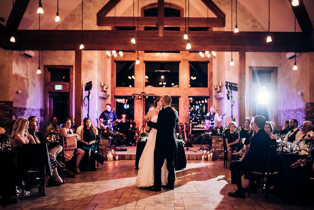 Wedding couple dancing in rustic venue with exposed beams and hanging lights.