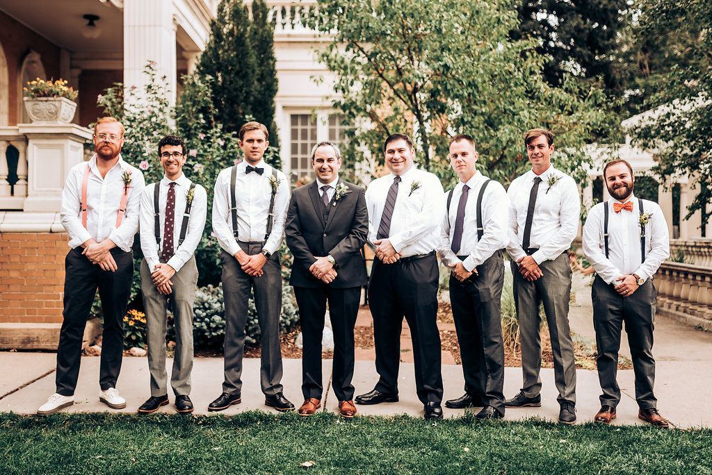 Groomsmen in suspenders and ties pose in a line outside a brick building.