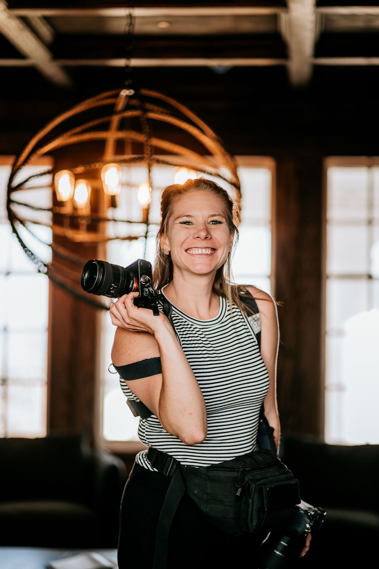 Portrait of a photographer in a striped tank top holding professional camera equipment in a rustic indoor setting.