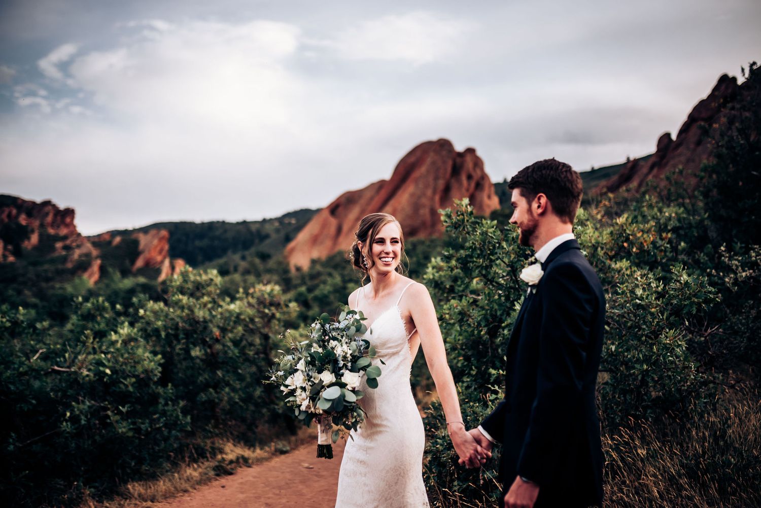 Couple in wedding attire poses on dirt path with dramatic red rock formations in background.