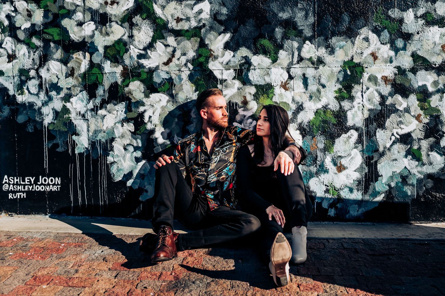 A couple posing against a floral backdrop wall in an urban setting.