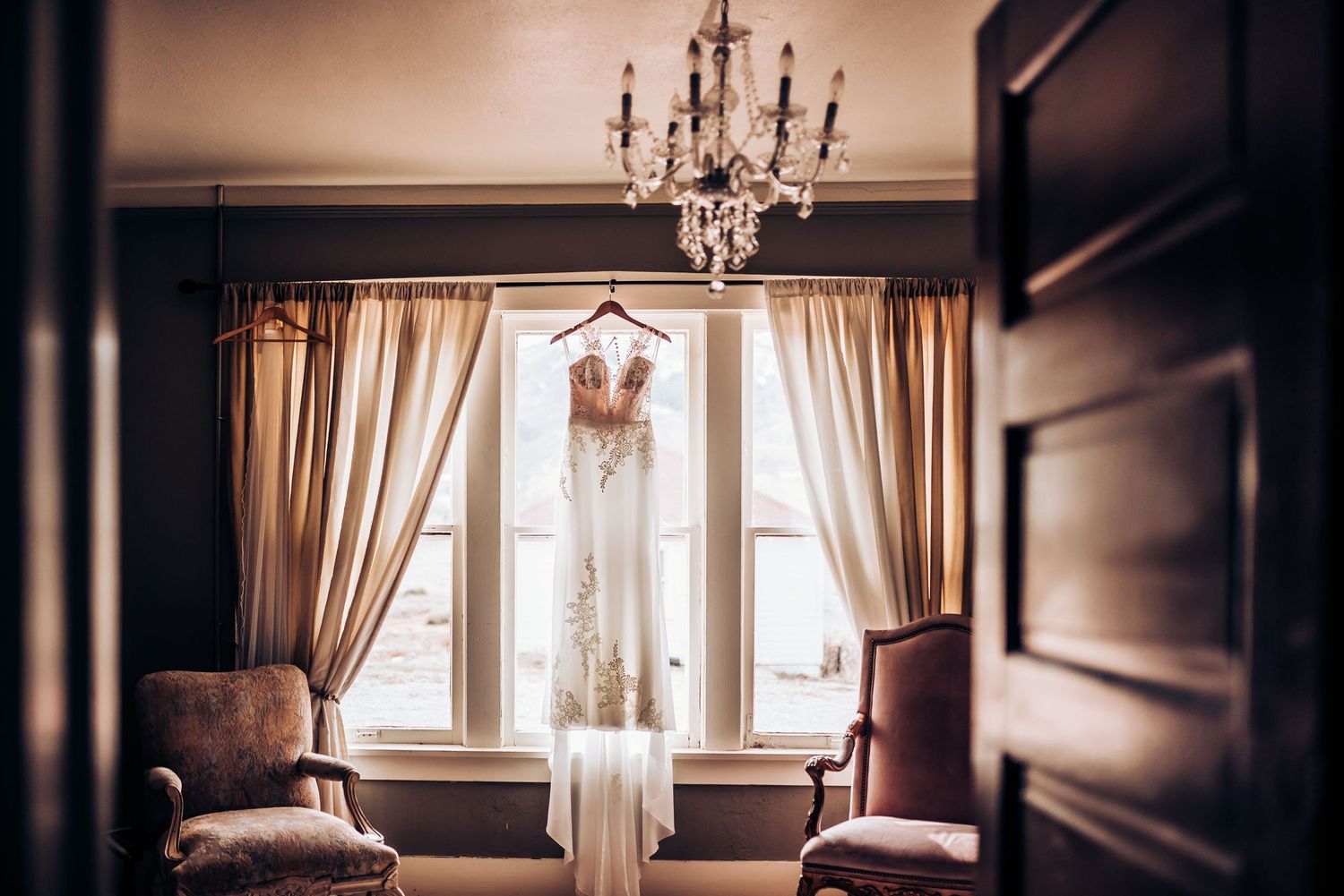 An elegant wedding dress hanging in a window frame between two chairs beneath a crystal chandelier.