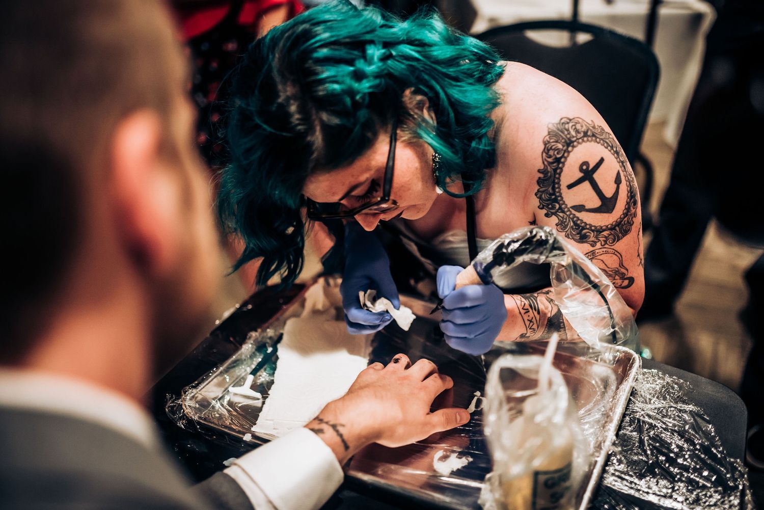 Tattoo artist with teal hair working on client during formal event.