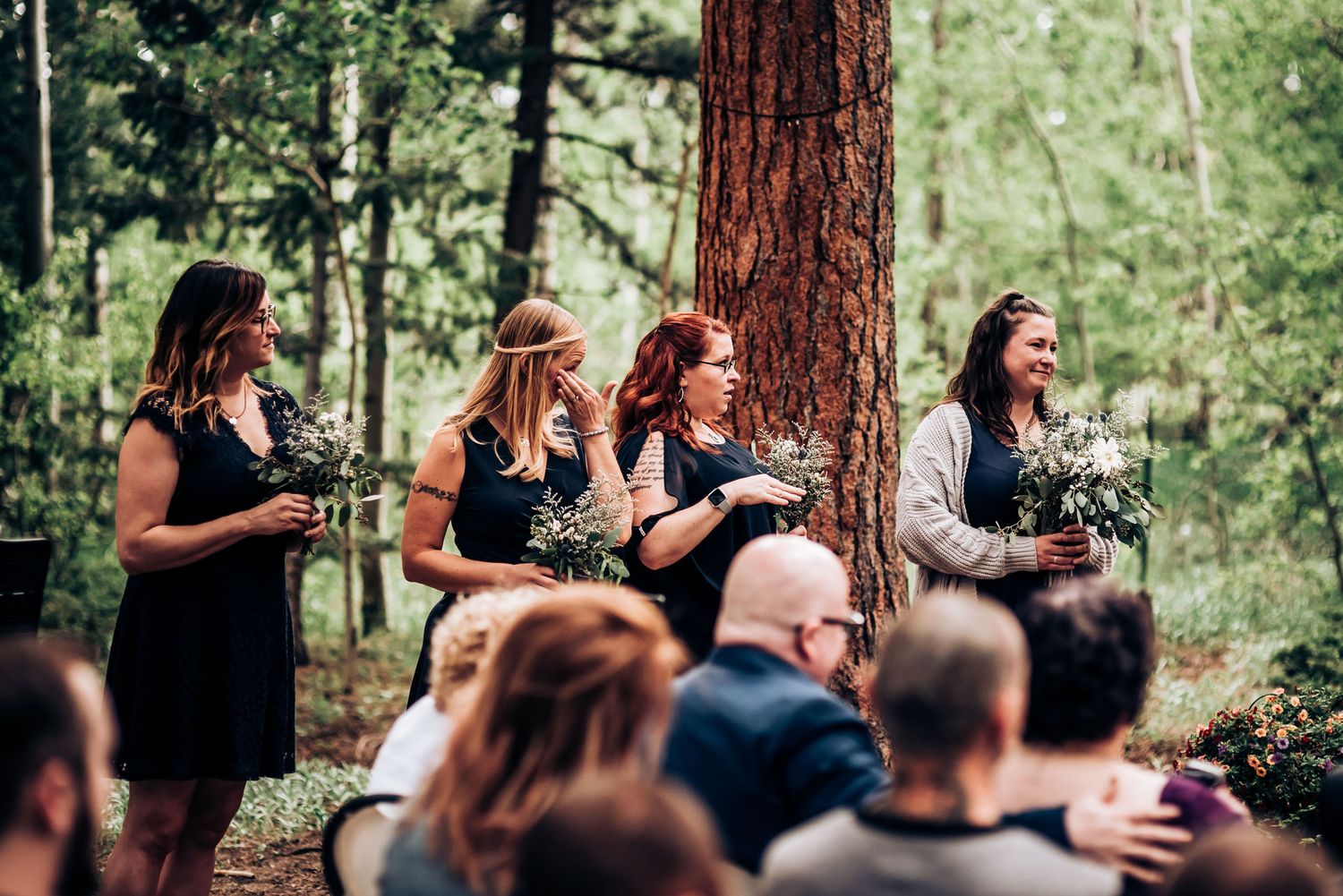 Wedding guests in black dresses standing near pine trees during an outdoor forest ceremony.