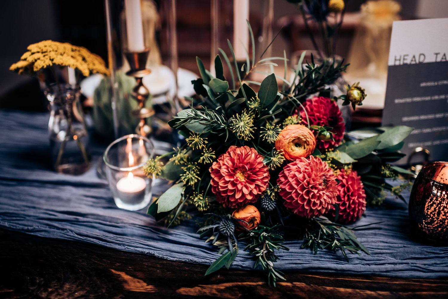 Vibrant floral centerpiece with red dahlias and greenery on dark tablecloth with candle accent.