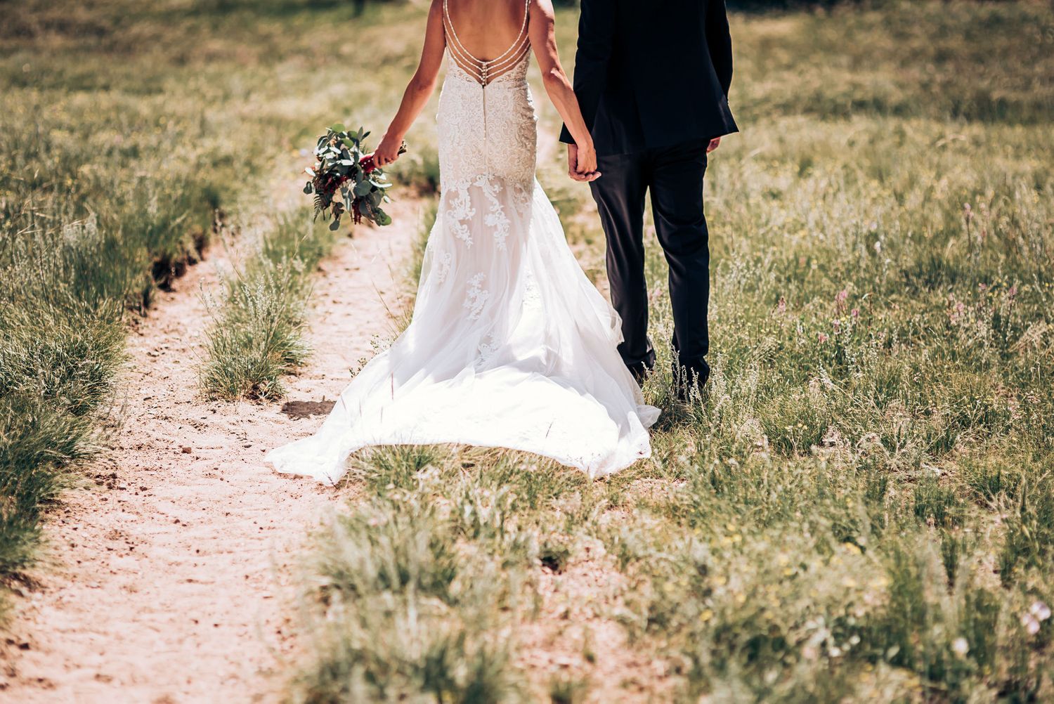 A close-up view of a bride and groom walking on a dirt path through golden grass fields.