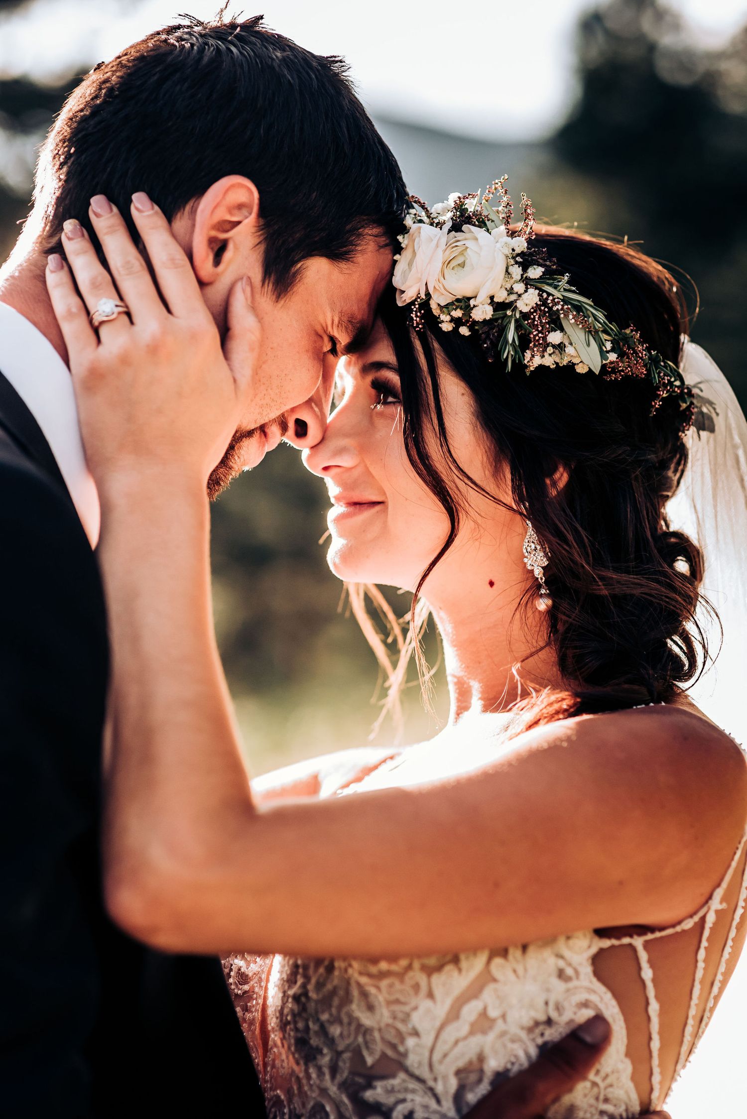 Sun-kissed wedding portrait shows tender moment with floral crown and lace dress details.