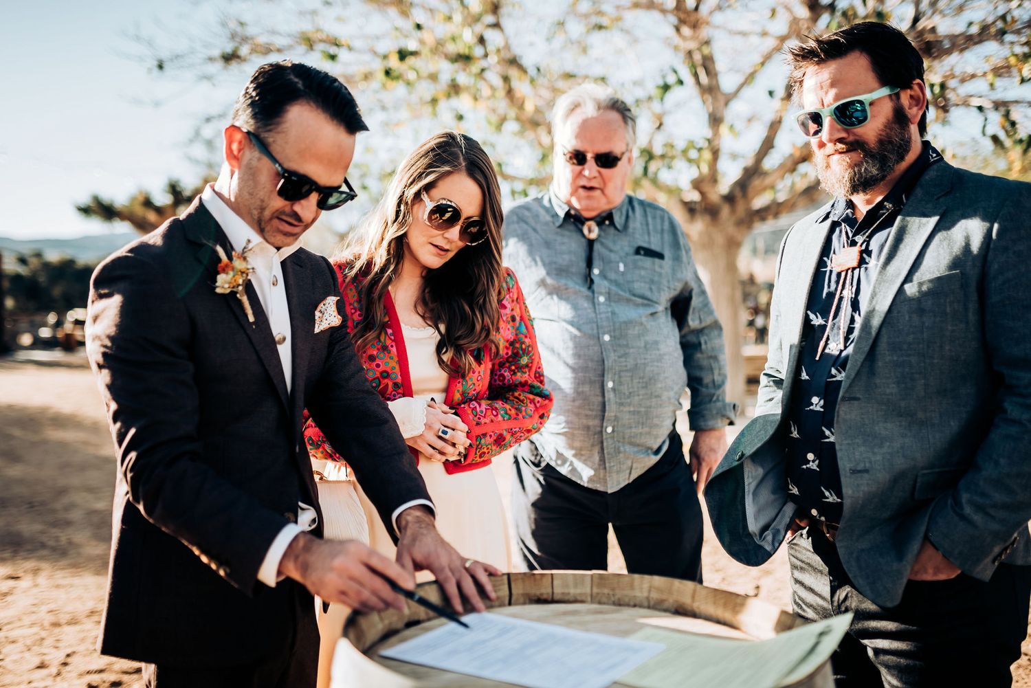 Group gathered around wooden barrel signing documents at outdoor desert celebration.