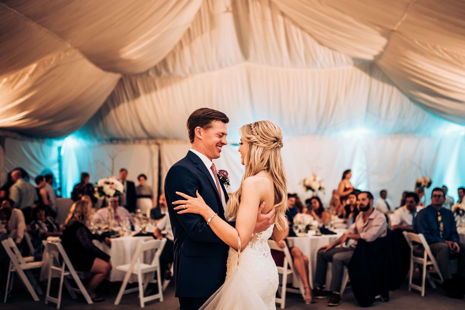 Wedding couple dances surrounded by guests seated at round tables in an elegant tented reception.