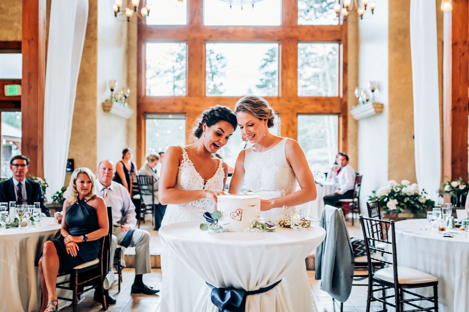 Wedding reception moment with two people in white dresses laughing while cutting cake together.